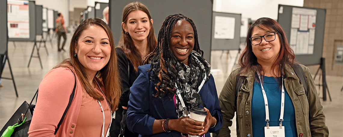 Four women smile at the camera in a conference setting with posters in the background.