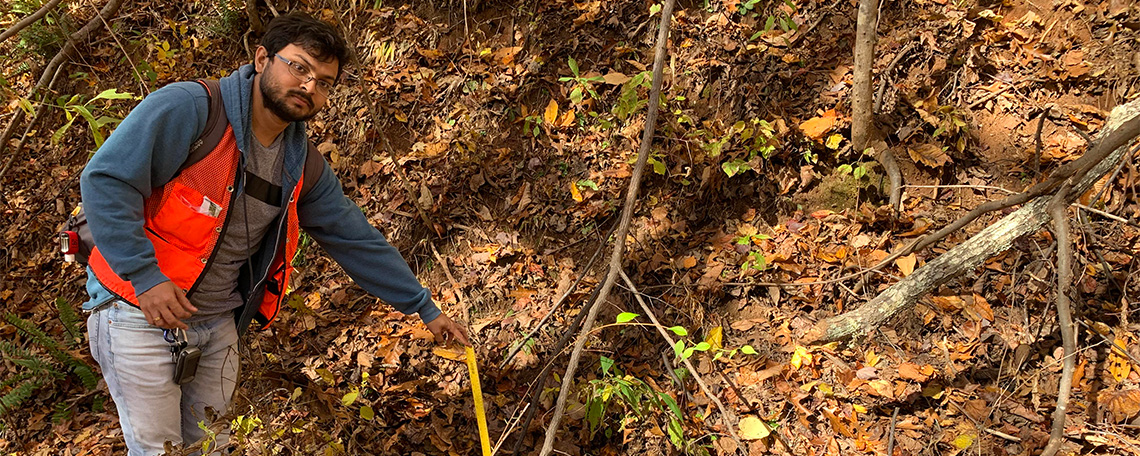 A man takes measurements on a hillside covered in fallen leaves.